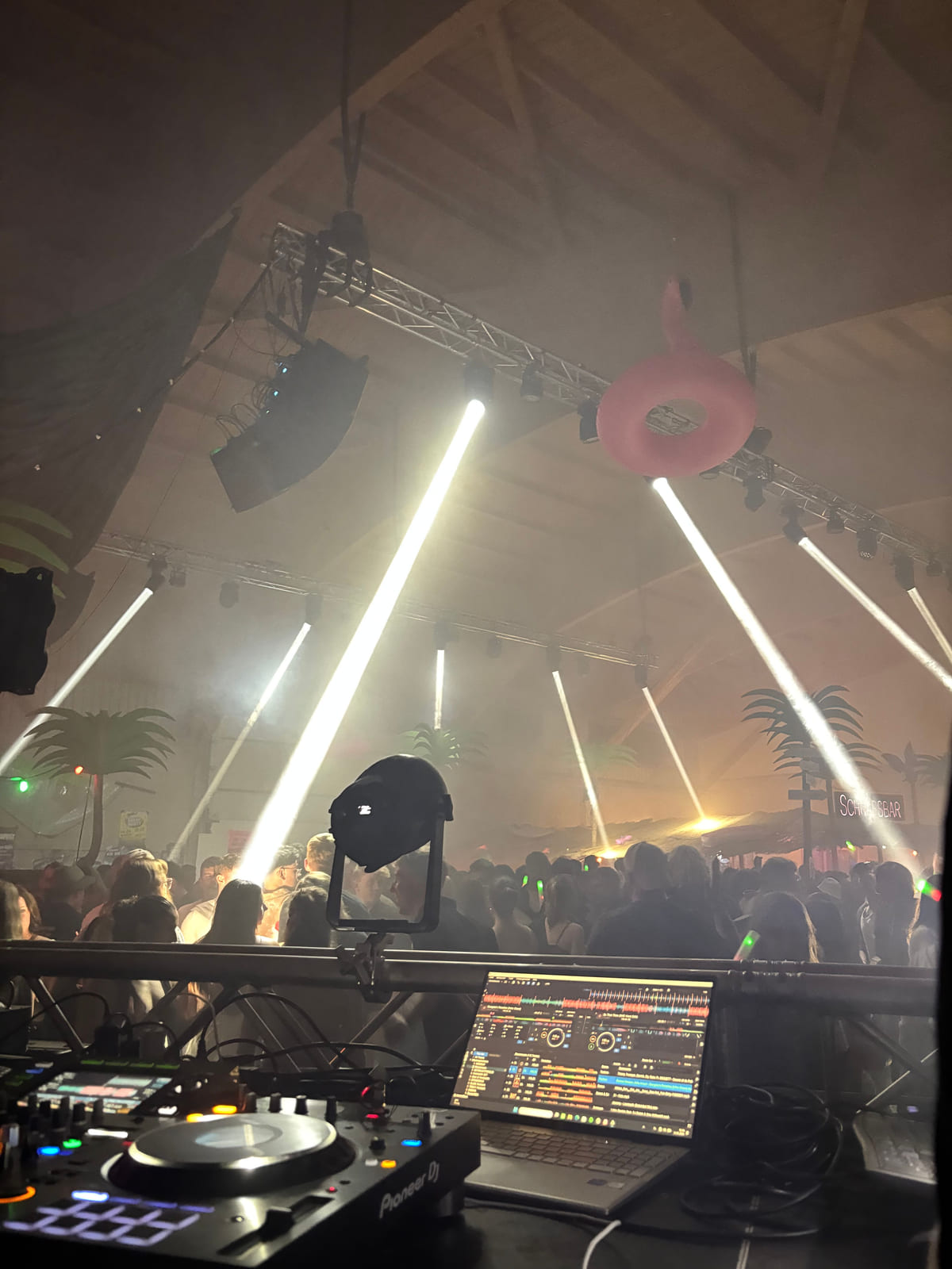Blick vom DJ-Pult in eine Halle mit Lichtstrahlen und Publikum